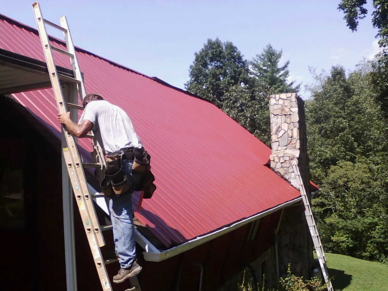 Roofer on a ladder beside a red metal roof and stone chimney — Soffit & Fascia Repair by Roofing Brothers in Britton